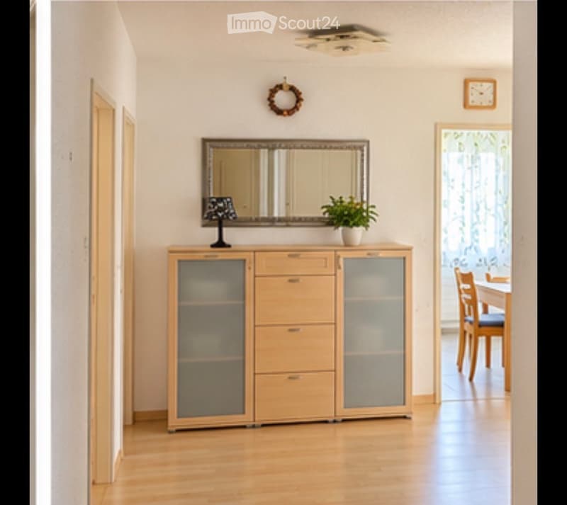 Bright hallway with wooden flooring, a cabinet with glass doors, a small table lamp, a potted plant, and a mirror above the cabinet. A doorway leads to a dining area with a table and chairs.
