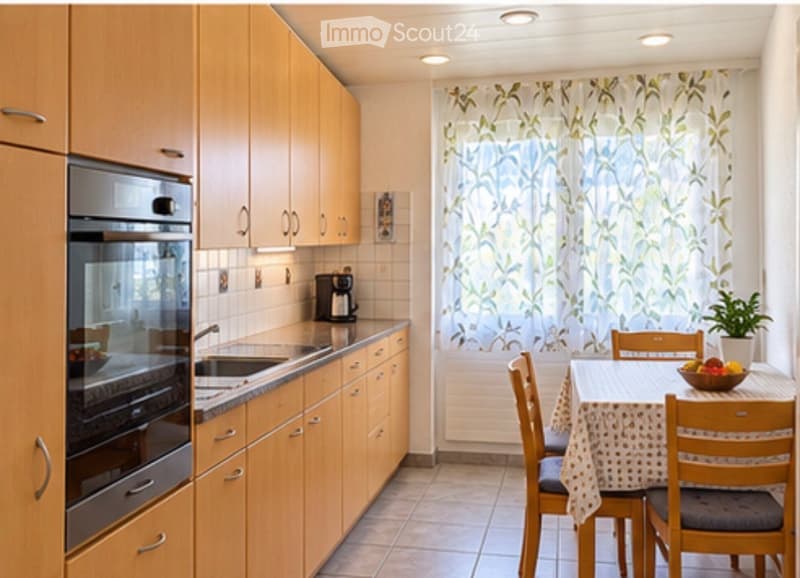 A modern kitchen with wooden cabinets, stainless steel oven, sink, and coffee maker. Adjacent dining area with a table, chairs, and a bowl of fruit on the table.