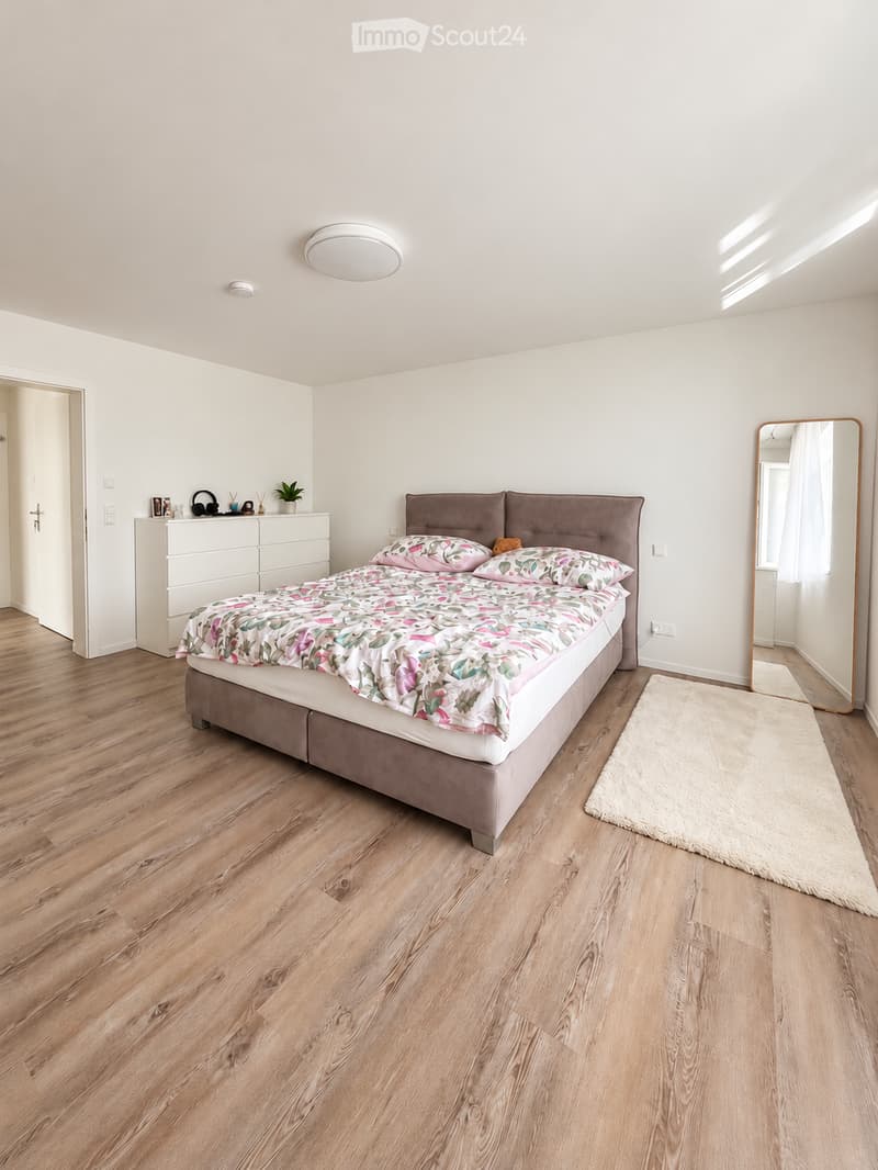 Bright bedroom with wooden flooring, a large bed with a floral duvet, a nightstand with a plant, and a full-length mirror on the wall.