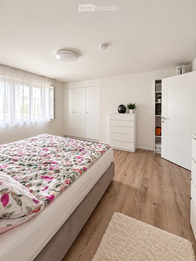 Bright bedroom with wooden flooring, a bed with a floral duvet, a white wardrobe, a dresser with a plant on top, and a white shaggy rug on the floor.