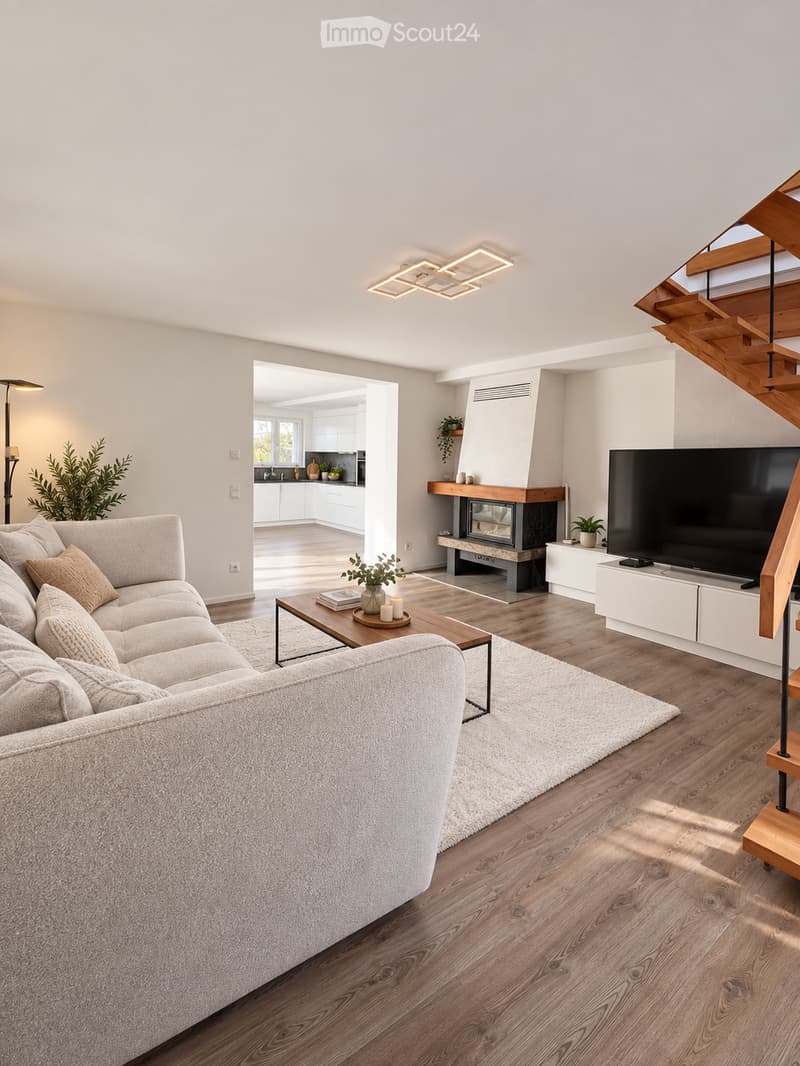 Modern living room with a large sectional sofa, coffee table, fireplace, and television. The space features wooden flooring, recessed lighting, and a view into an adjacent kitchen area.