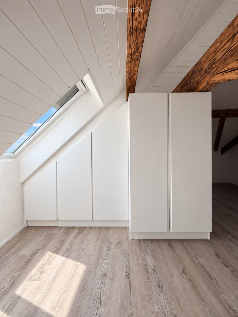 Bright, empty attic space with wooden flooring, white paneled walls, and exposed wooden beams. Skylight windows allow natural light to fill the room.