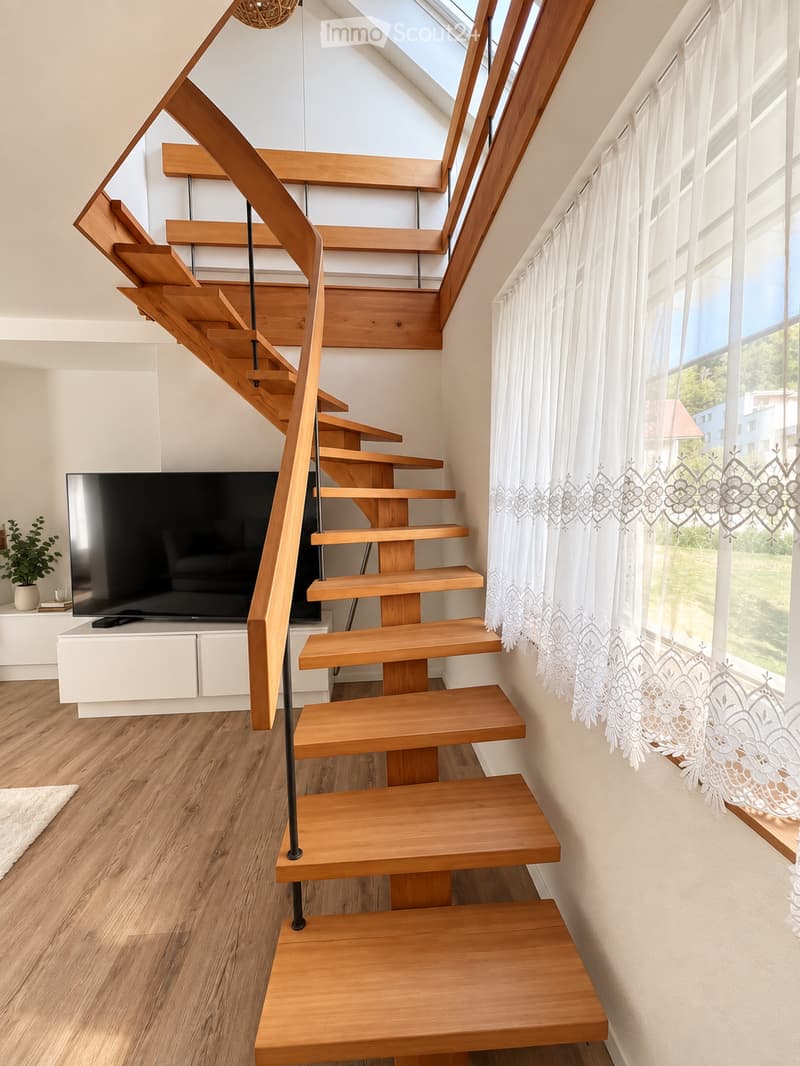Modern living room with a wooden staircase leading upwards, large flat-screen TV on a white stand, a small plant on a table, and white curtains on large windows.