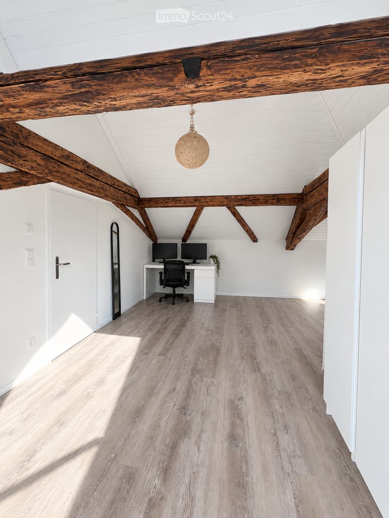 Bright, airy room with exposed wooden beams, white walls, light wooden flooring, and a modern desk setup with two computer monitors and a chair.