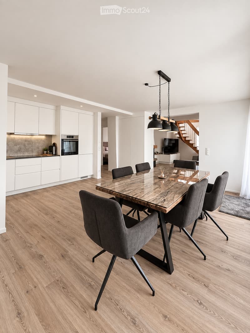 Modern dining area with a wooden dining table and grey chairs, adjacent to a white kitchen with cabinets and an oven. A staircase and living area with a TV are visible in the background.