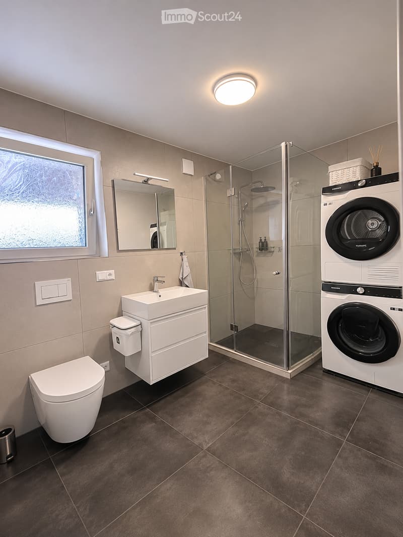Modern bathroom with a toilet, sink, mirror, shower, washing machine, and dryer. Features a large window, light fixture, and dark tiled floor.