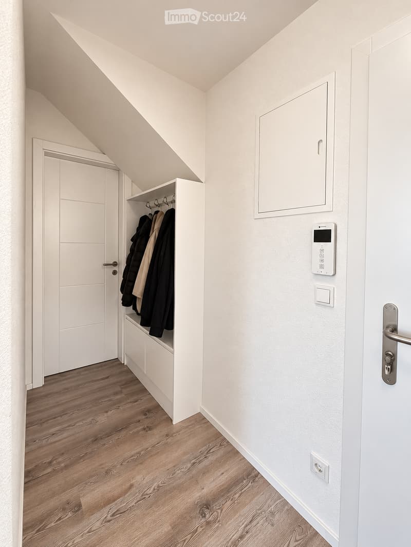 A narrow hallway with wooden flooring, white walls, built-in closet with coats, and a door with a modern handle.