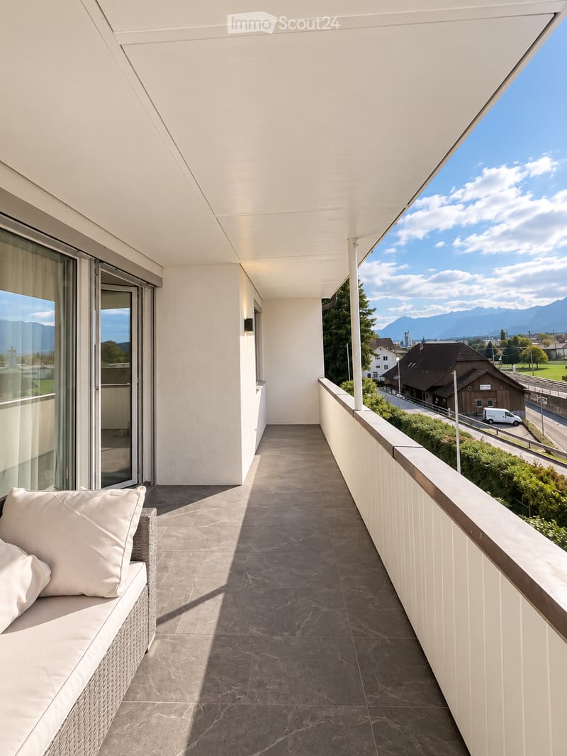 Balcony with grey tiled floor, white wicker sofa with cushions, large windows, and scenic mountain view.