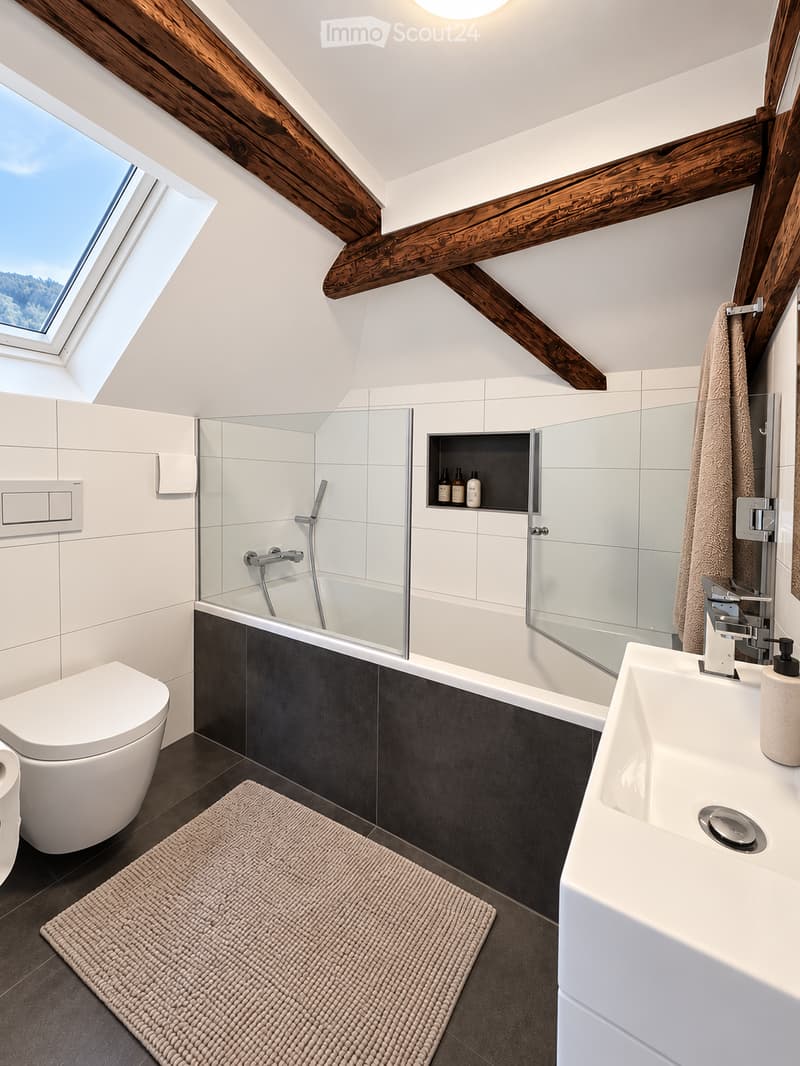 Modern bathroom with a skylight, white tiles, dark grey accents, a bathtub with a glass enclosure, a toilet, a sink, and a built-in shelf holding bottles.