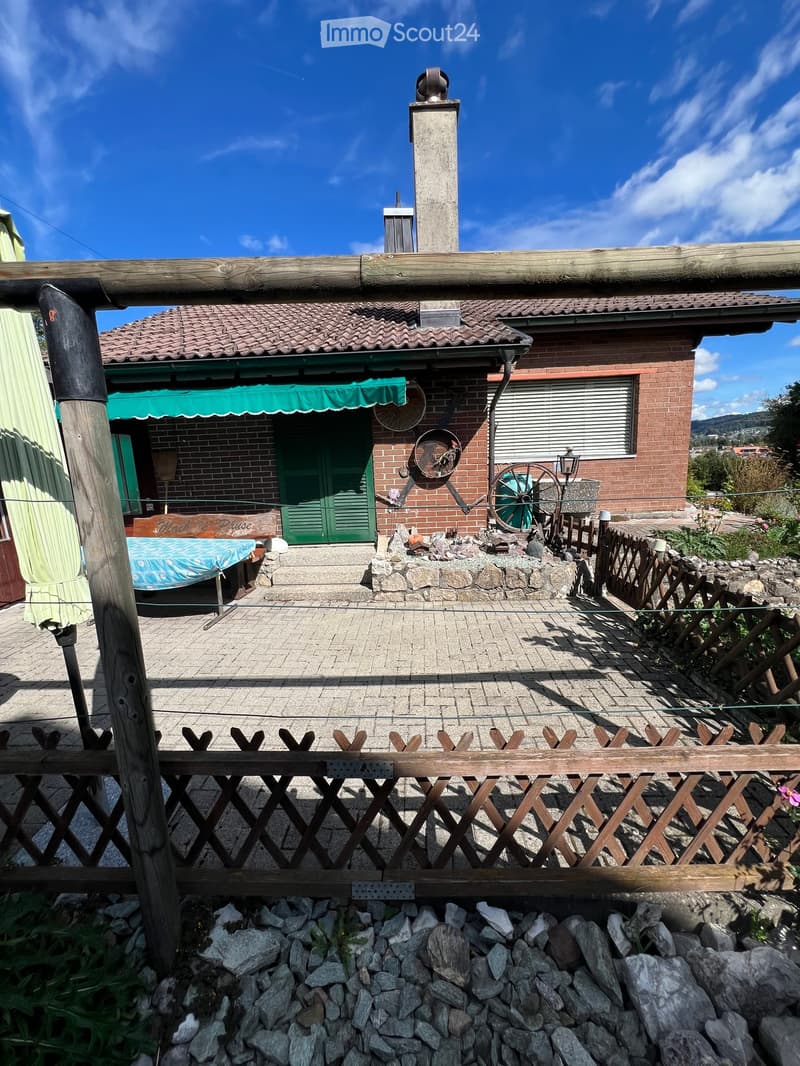A two-story brick house with a tiled roof, green shutters, and a chimney. The entrance features a green awning and a small porch with a table and chairs. A wooden fence and stone wall surround the property.