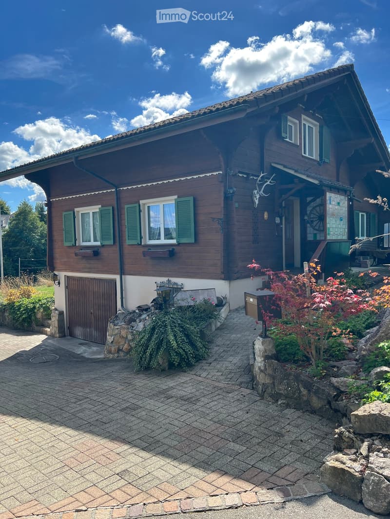 Two-story wooden house with green shutters, a garage, and a well-maintained garden with stone pathways and decorative plants.