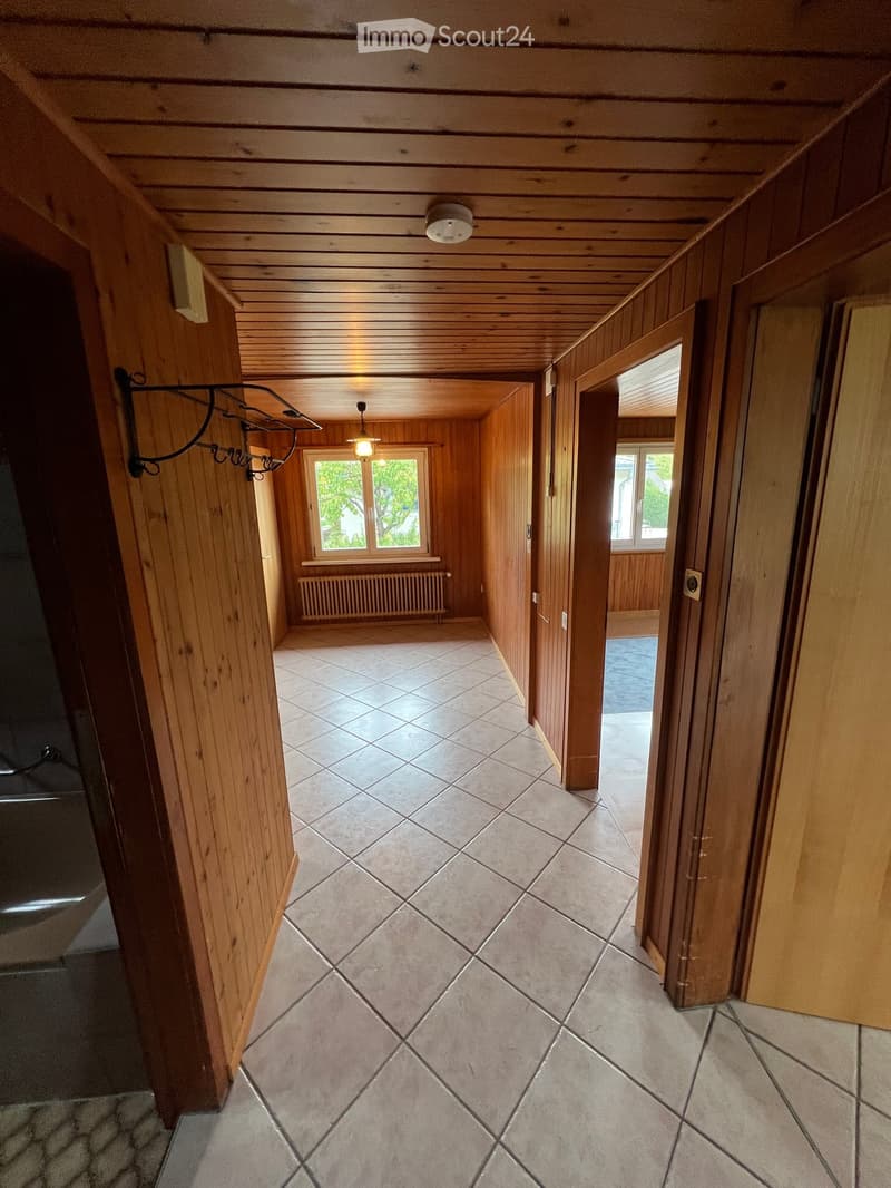 Wooden-paneled hallway with tiled floor, ceiling lights, and a coat rack on the left side. A window and radiator are visible in the background.
