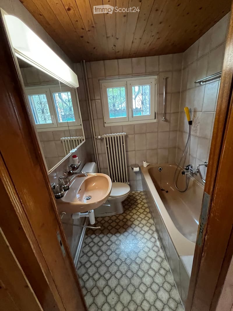 Bathroom with beige tiles, a sink, toilet, bathtub, radiator, and a mirror above the sink. The room has wooden beams on the ceiling and a window with decorative bars.