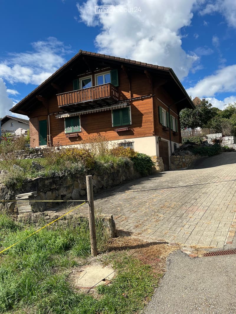 Two-story wooden house with green shutters, balcony, stone pathway, and garden area.
