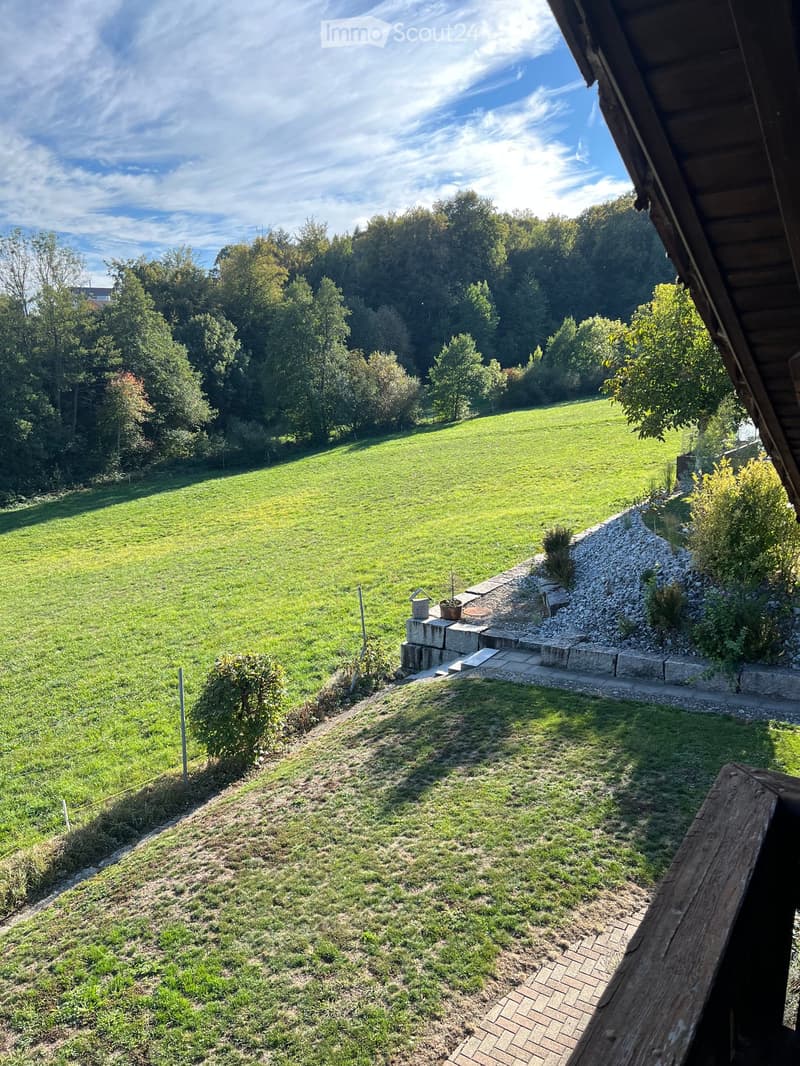 A spacious, well-maintained garden with a large grassy area, a gravel pathway, and a small stone platform with a small plant and a bucket. The garden is bordered by trees and shrubs, with a wooden deck visible in the foreground.