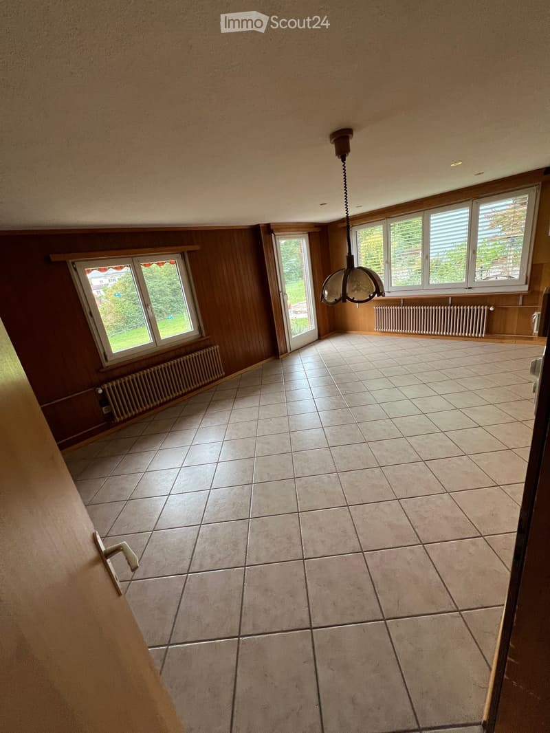 Empty room with tiled floor, wooden walls, large windows, radiator, and a hanging light fixture.