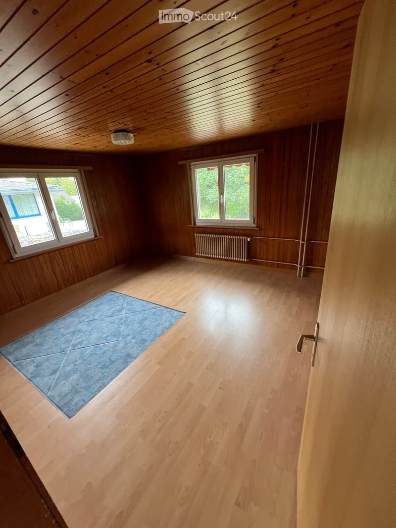 Empty room with wooden walls and floor, two windows, a radiator, and a small blue mat on the floor.