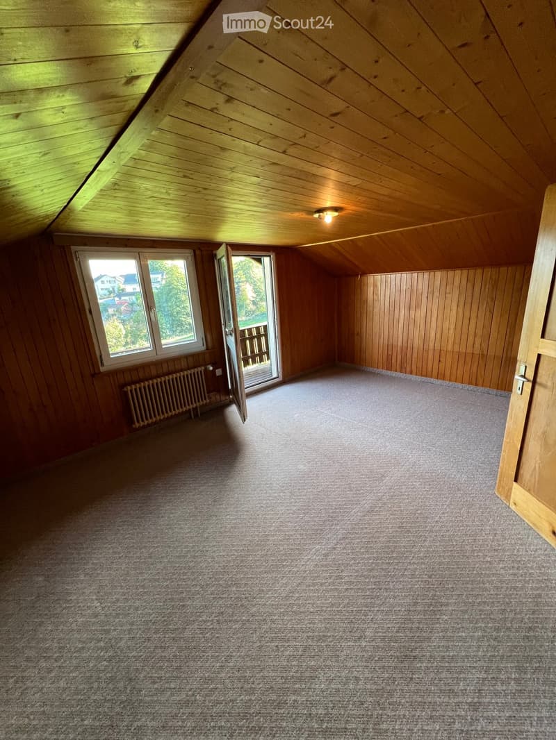 Empty attic room with wooden walls and ceiling, carpeted floor, large window, open door leading to a balcony, and a radiator.