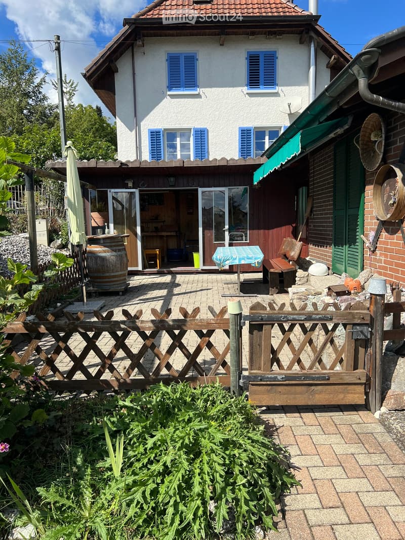 A cozy outdoor area with a wooden fence, a patio, and a small seating area with a table and chairs. The house has a white exterior with blue shutters and a green awning over the entrance.