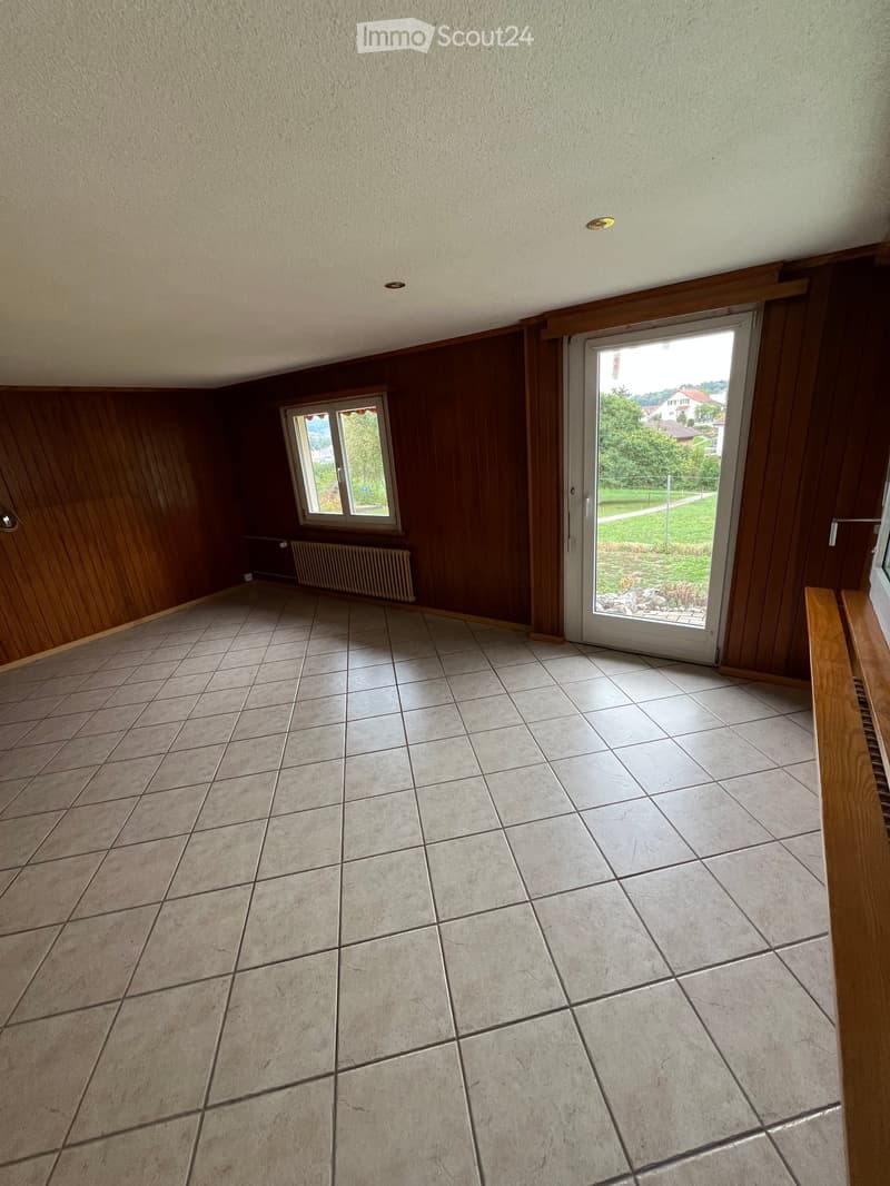 Empty room with tiled floor, wooden walls, and a large sliding glass door leading to a green outdoor area.