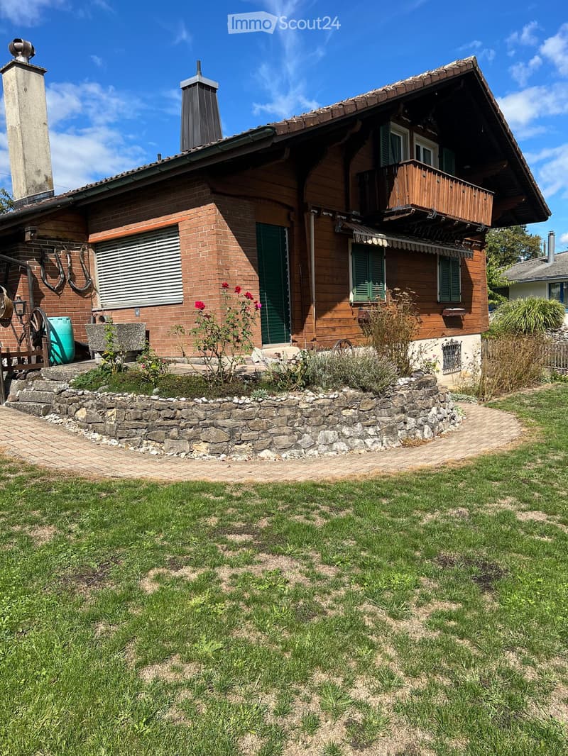Two-story house with brick and wooden exterior, green shutters, balcony, stone pathway, and garden area.