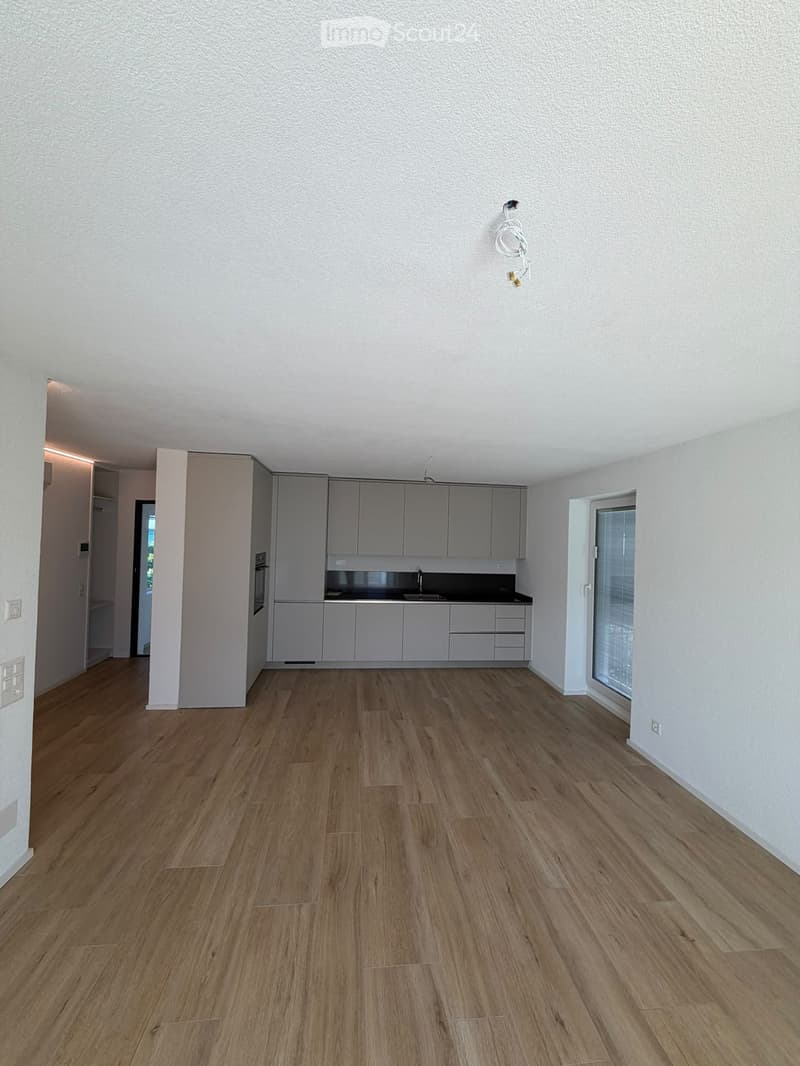Empty kitchen with light grey cabinets, wooden flooring, and a window with blinds. No appliances or furniture are present.