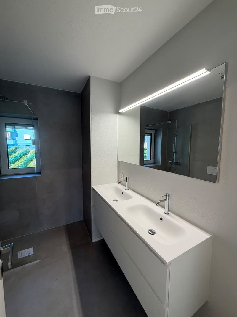 Modern bathroom with a double sink, sleek white cabinetry, and large mirror with built-in lighting. The space features a window with a view of greenery outside.