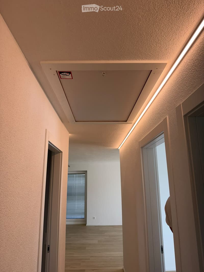 Empty hallway with light wooden flooring, white walls, and recessed lighting. Doors lead to adjacent rooms, one with a window and blinds visible.