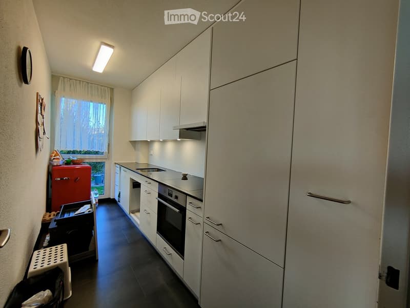 Modern kitchen with white cabinets, stainless steel appliances, and a red refrigerator. Features a window with a view of greenery, a sink, and a built-in oven.