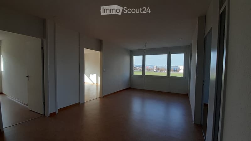 Empty hallway with wooden flooring, white walls, and large windows showing an outdoor view. No furniture or equipment present.