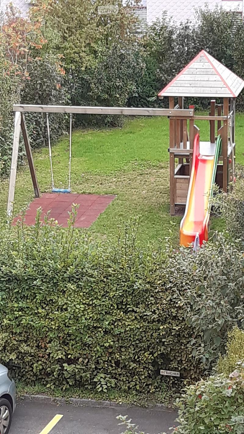 A small, enclosed garden area with a wooden play structure featuring a slide and swing, surrounded by dense green hedges and a paved pathway.
