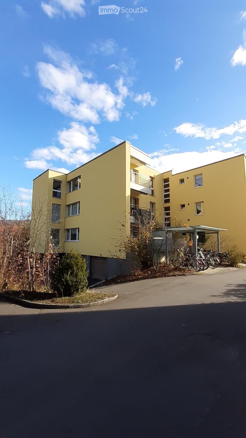A modern yellow apartment building with multiple windows and balconies, surrounded by a paved area with bicycles and some greenery.
