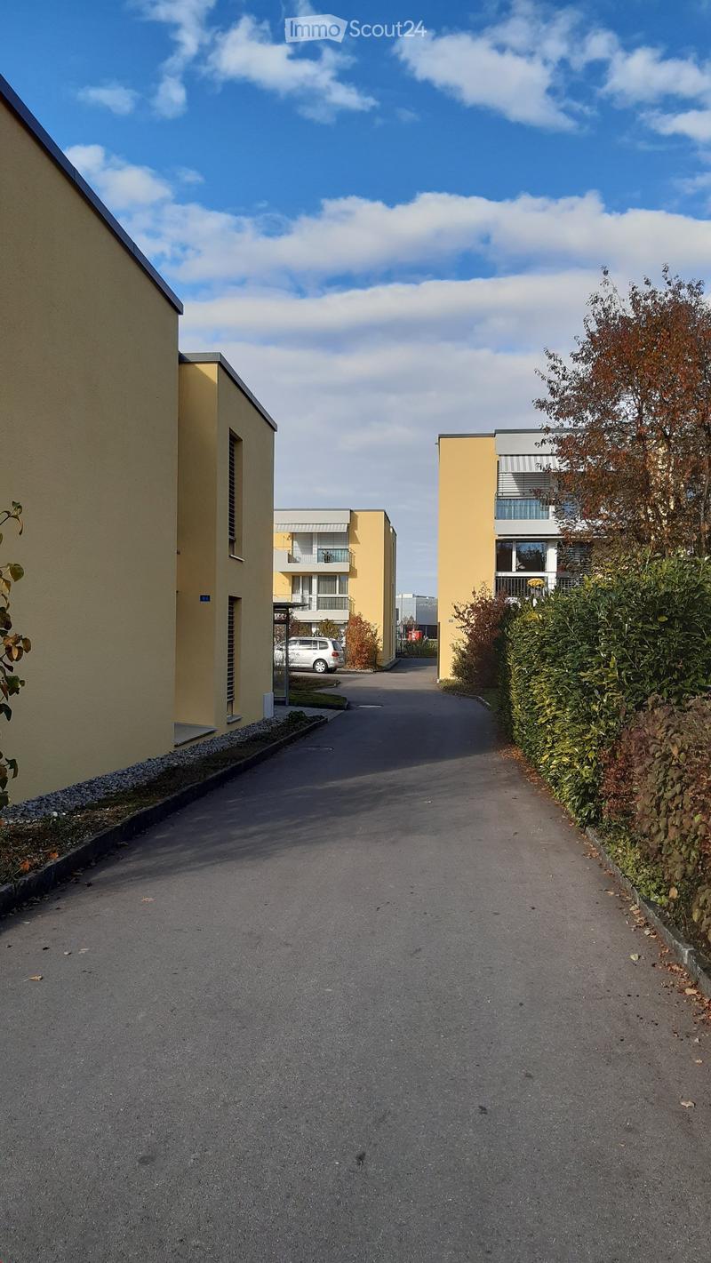 A paved pathway between two yellow buildings with balconies, surrounded by greenery and a parked car in the background.
