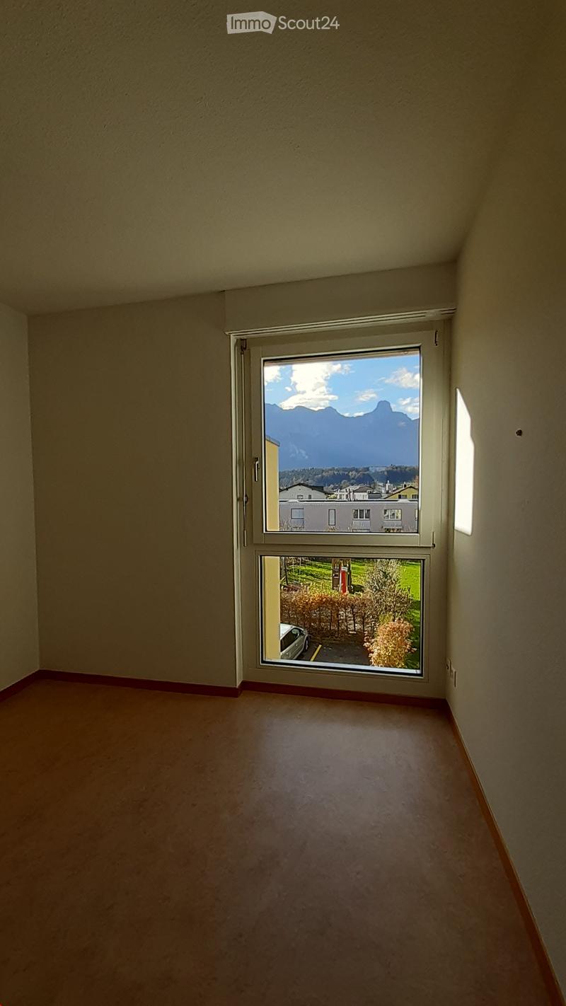 Empty room with beige walls and carpeted floor, featuring a large window with a view of a landscape and a parking area.