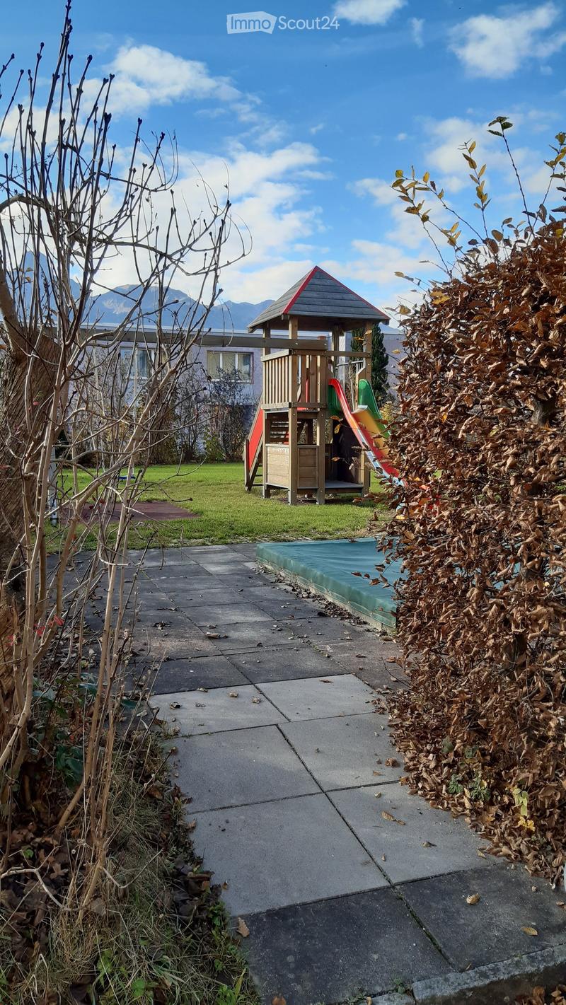 Outdoor area with a small playground structure, slide, and a covered playhouse. The area is surrounded by grass, trees, and a paved walkway.