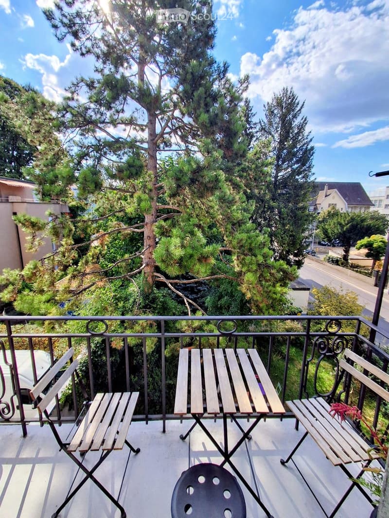 Balcony with a small table and three chairs, overlooking a green, tree-filled area with houses in the background.
