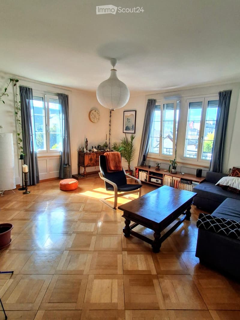 Bright living room with wooden flooring, large windows, a sofa, coffee table, armchair, and decorative plants.