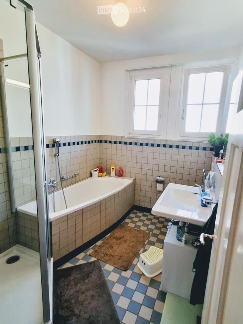 Bathroom with a bathtub, sink, shower, and a small stool. The room features tiled walls and a checkered floor with a small brown rug.