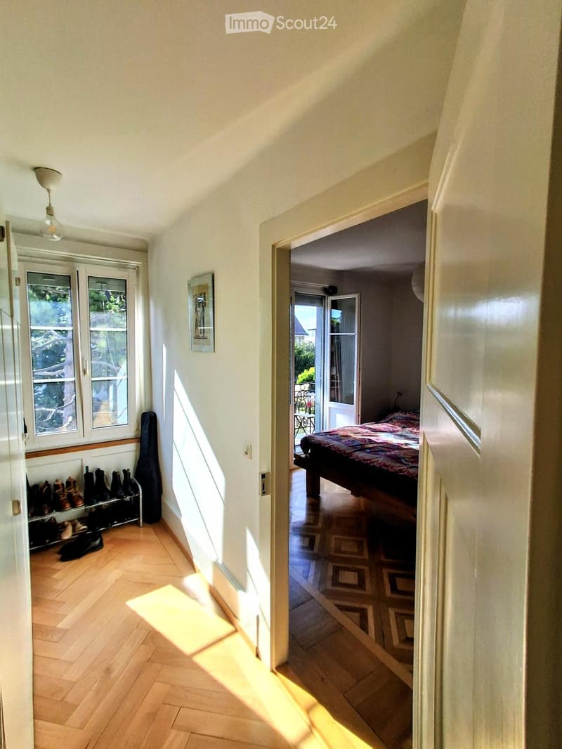 Bright hallway with wooden flooring, white walls, and a ceiling light fixture. A shoe rack with several pairs of shoes is visible on the left, and an open door leads to a bedroom with a bed and patterned bedding.