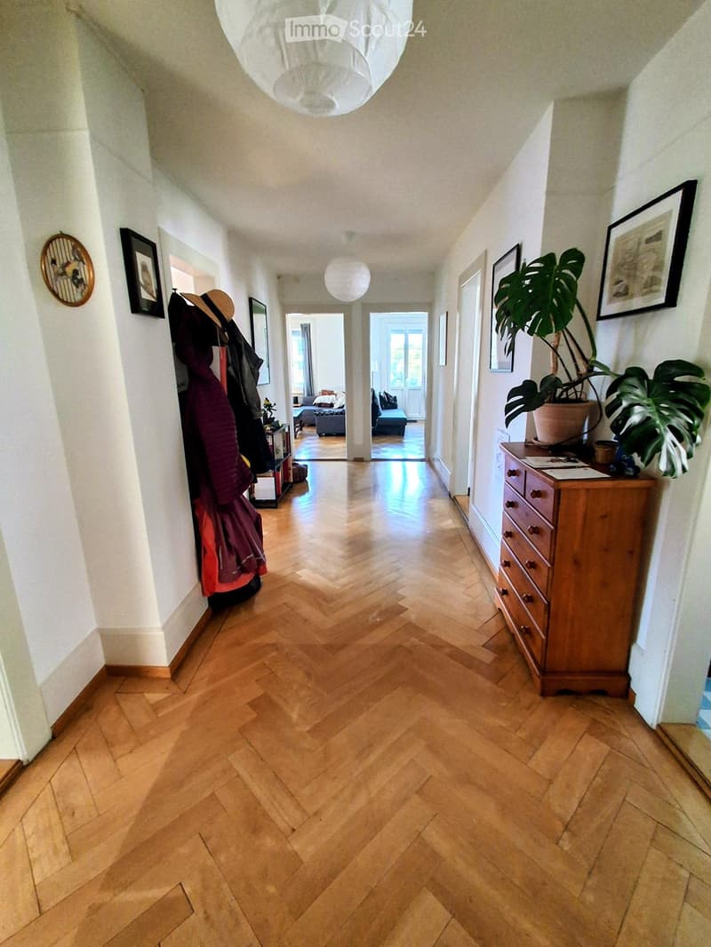 A long, well-lit hallway with herringbone wooden flooring, white walls, and several framed pictures. A coat rack with jackets and a potted plant are visible on either side, leading to adjacent rooms.