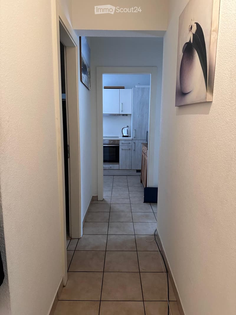 A narrow hallway with tiled flooring, white walls, and a view into a small kitchen area with white cabinets and a kettle on the counter.