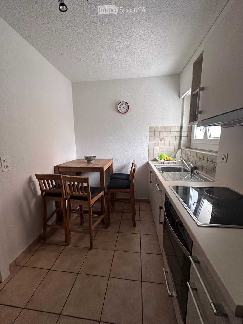 Compact kitchen with tiled floor, white walls, and modern appliances including a sink, oven, and countertop. A small wooden table with chairs is positioned near the wall, and a clock is mounted on the wall.