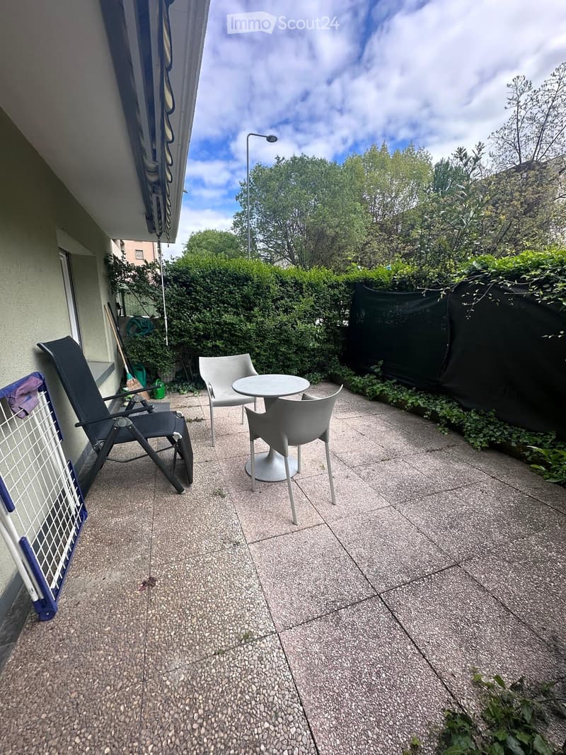 Balcony with a small round table and two chairs, surrounded by greenery and a black privacy screen.