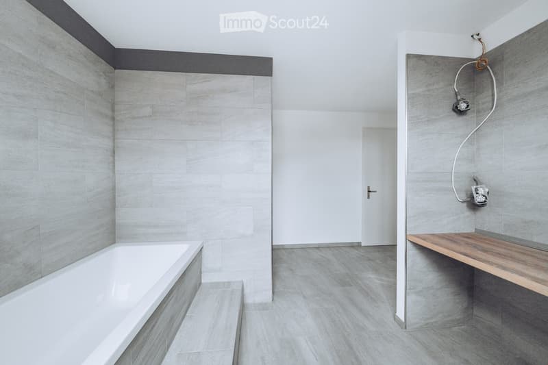 Modern bathroom with a white bathtub, grey tiled walls, and a wooden shelf with a showerhead and electrical outlet.
