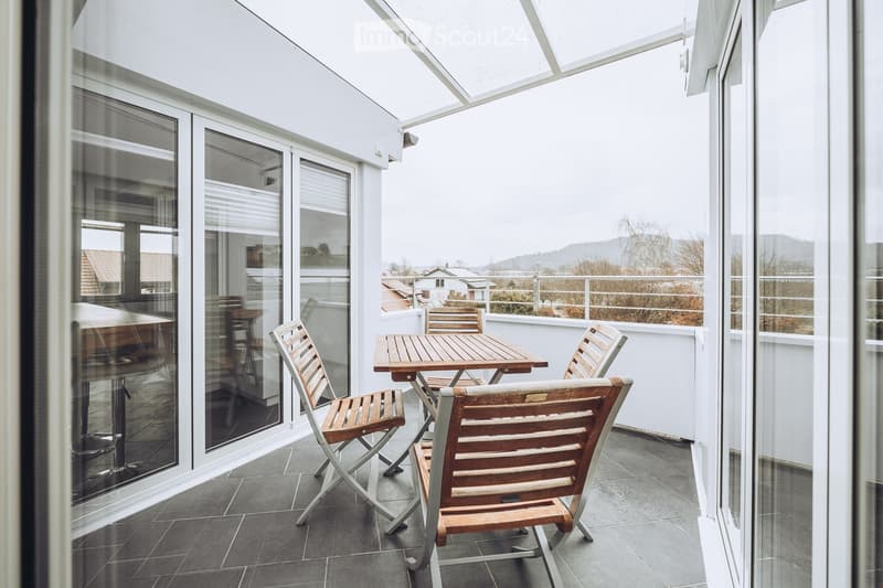 Balcony with a wooden table and chairs, large glass windows, tiled floor, and a view of the surrounding area.