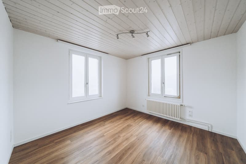 Empty room with white walls, wooden floor, two windows, radiator, and track lighting fixture on the ceiling.