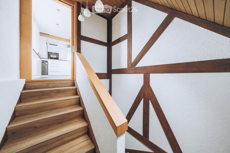 Wooden staircase with a handrail, white walls, and exposed wooden beams on the ceiling. The background shows a glimpse of a modern kitchen area with white cabinets and stainless steel appliances.