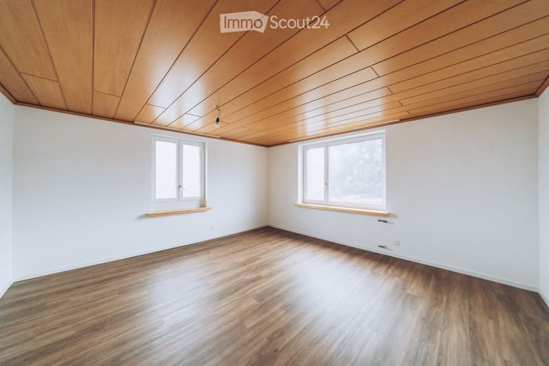 Empty room with wooden flooring, white walls, and a wooden ceiling. Two windows with white frames and wooden sills are present.