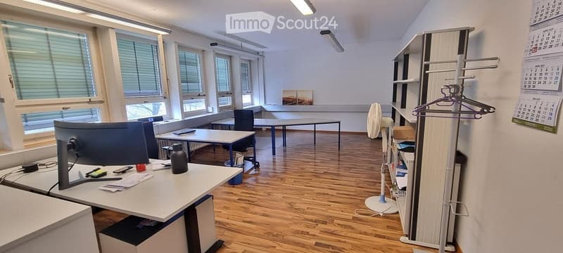 An office room with wooden floors, several desks and chairs, a monitor, office items, a cabinet, a fan, and a calendar on the wall.