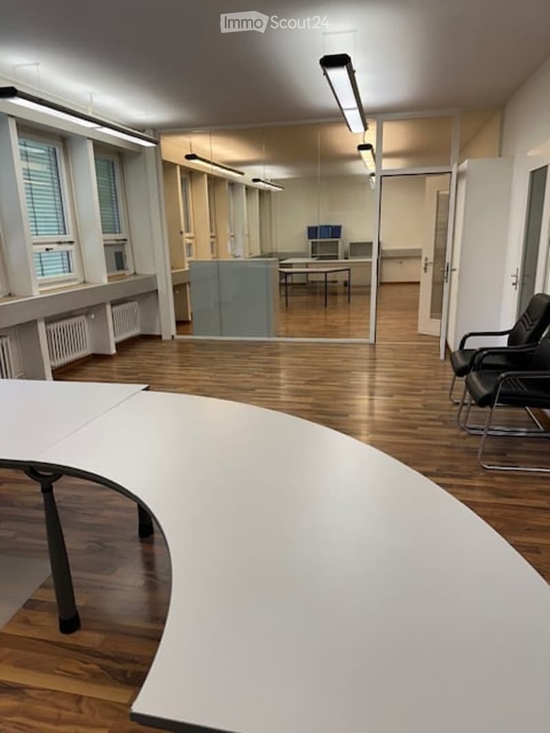 Modern office space with wooden flooring, large windows, white curved table, black chairs, and glass partition.